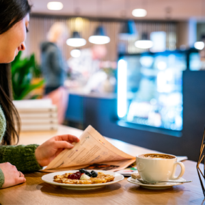 Kvinne som leser avisen med vaffel og kaffekopp foran seg, i kafémiljø